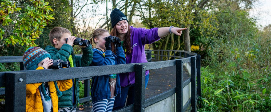 Ranger on boardwalk pointing whilst children with binoculars look